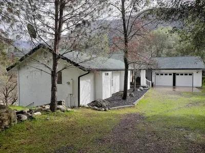 a white house with two garage doors and trees at Sierra River House 5 min to Sequoia Park Entrance in Three Rivers