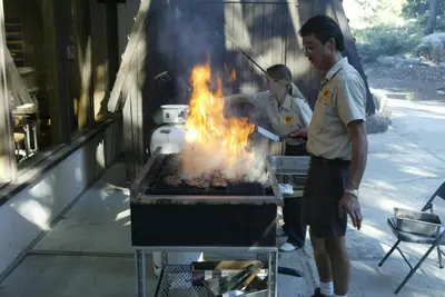 two people cooking food on a grill with flames at Snowflower Camping Resort Cottage 7 in Emigrant Gap